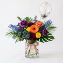 The Birthday Pop colorful floral arrangement with orange, purple, yellow and burgundy flowers and a clear confetti balloon.