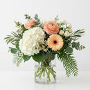 Radiant Celebration Bouquet with white hydrangea, soft peach flowers and layered greenery in a clear glass vase.