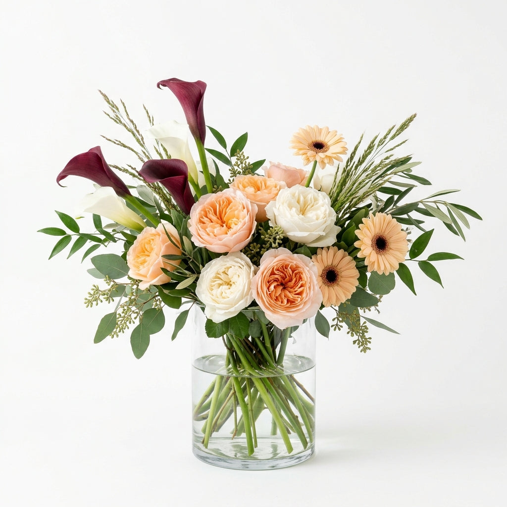 Tropical Birthday Chic arrangement with peach and cream roses, burgundy calla lilies and peach gerbera daisies in a glass vase.