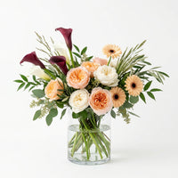 Tropical Birthday Chic arrangement with peach and cream roses, burgundy calla lilies and peach gerbera daisies in a glass vase.