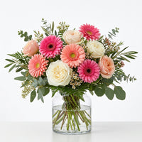 Blush & Celebration pink gerbera daisies with blush and cream roses and greenery arranged in a clear glass vase.