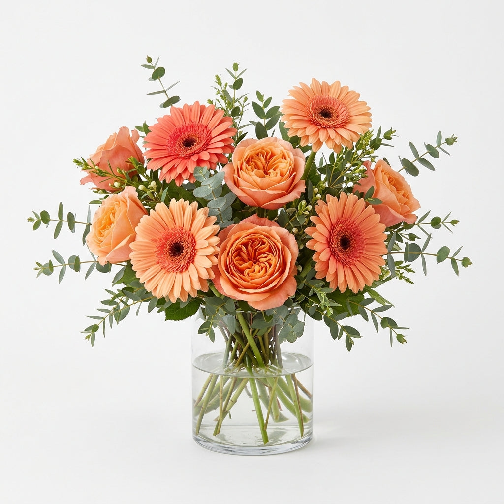 The Miami Birthday Standard arrangement with peach and coral roses, gerbera daisies and greenery in a clear glass vase.